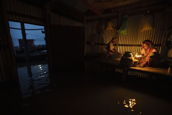 Yaad Ali, 55, left, offers prayers as his wife Monuwara Begum, 45, cooks food in their submerged house in Sandahkhaiti, a floating island village in the Brahmaputra River in Morigaon district, Assam, India, Tuesday, Aug. 29, 2023. (AP Photo/Anupam Nath)