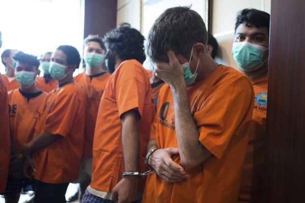Un australiano, identificado únicamente como P.R. (delante, derecha), y que está esposado a un ciudadano indio indentificado como H.V., reacciona durante su presentación a la prensa junto a otros detenidos por presunto narcotráfico, durante una conferencia de prensa en Denpasar, Bali, Indonesia, el 5 de junio de 2025. (AP Foto/Firdia Lisnawati)