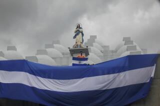 FILE - A masked youth holds a Nicaraguan national flag while standing on the roof of the Cathedral during a one year memorial marking a government crackdown on a Mother's Day march in Managua, Nicaragua, May 30, 2019. Nicaragua’s police have prohibited a religious procession to the Cathedral scheduled for Saturday, Aug. 13, 2022. (AP Photo/Alfredo Zuniga, File)