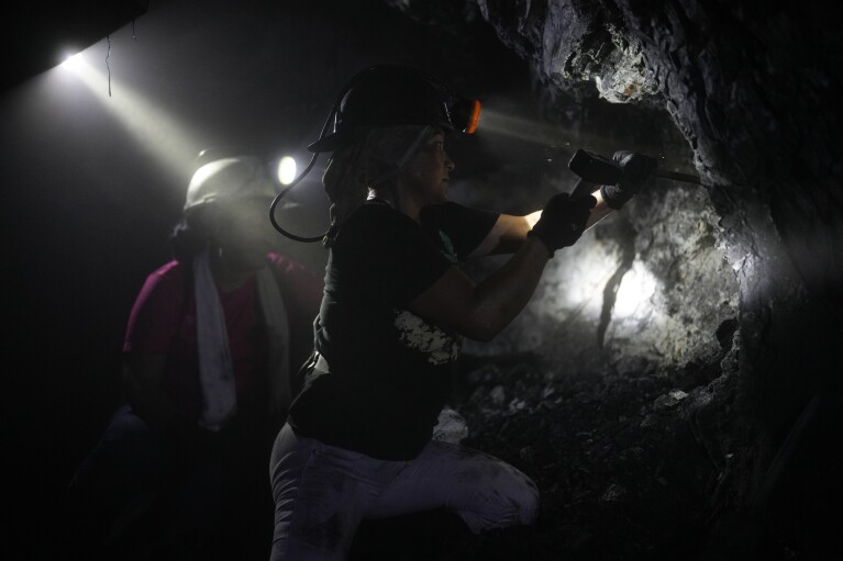 Wearing a headlamp, Margot Avila, 45, uses a hammer and chisel to loosen rocks from the tunnel of an informal mine near the town of Coscuez, Colombia, Wednesday, Feb. 28, 2024. The rocks that break off the walls are carried outside in carts, washed and sifted in hopes of finding emeralds. (AP Photo/Fernando Vergara)