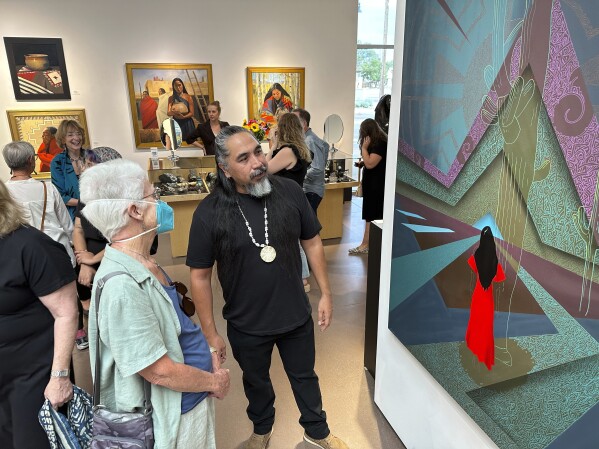 Artist Thomas "Breeze" Marcus attends an exhibition opening of his work at Blue Rain Gallery in Santa Fe, N.M. on Thursday, Aug. 17, 2023, including the painting titled "Answered Prayer," background. A separate group exhibition that included his work was postponed in Arizona amid outrage over a painting by Shepard Fairey that is critical of police. (AP Photo/Morgan Lee)