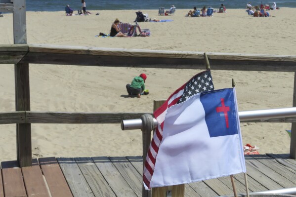 People sit on the beach on a weekday afternoon in Ocean Grove, N.J. on May 2, 2024 as a Christian flag and an American flag flutter in the breeze. The state of New Jersey says the Ocean Grove Camp Meeting Association is violating state beach access laws by keeping people off the beach until noon on Sundays. (AP Photo/Wayne Parry)