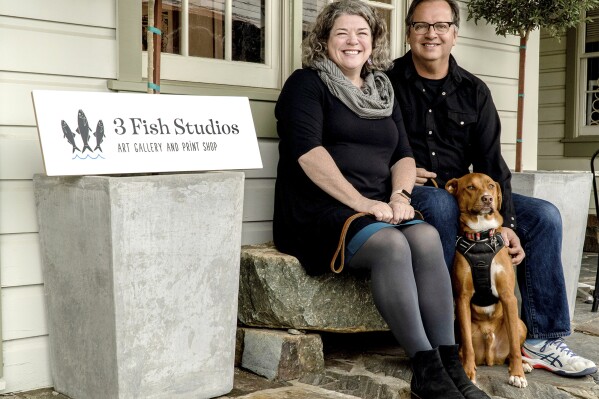 Husband and wife artist team Annie Galvin, left, and Eric Rewitzer pose with their dog Woody in front of their new 3 Fish Studios art gallery in Amador City, Calif., in 2023. Galvin and Rewitzer moved out of San Francisco to live closer to nature after their sabbatical in France and Ireland. (Eric Rewitzer via AP)