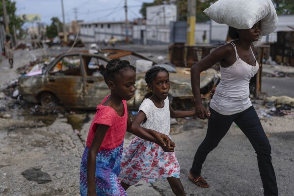 FILE - Girls holding hands are led past a burnt car blocking the street as they evacuate the Delmas 22 neighborhood to escape gang violence in Port-au-Prince, Haiti, May 2, 2024. As young Haitians are increasingly exposed to violence, the country is undergoing a wider push to dispel a long-standing taboo on seeking therapy and talking about mental health. (AP Photo/Ramon Espinosa, File)