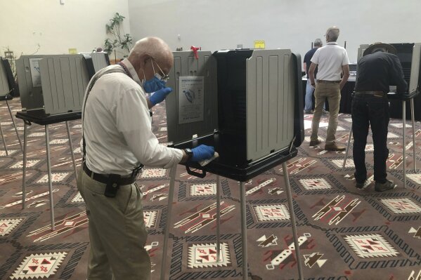 Temporary election worker Joseph Banar disinfects voting stations as a precaution against the coronavirus, on the first day of balloting in New Mexico at the Santa Fe Convention Center on Tuesday, Oct. 6, 2020, in Santa Fe. Election officials also are witnessing a massive surge in absentee ballot requests. (AP Photo/Morgan Lee)
