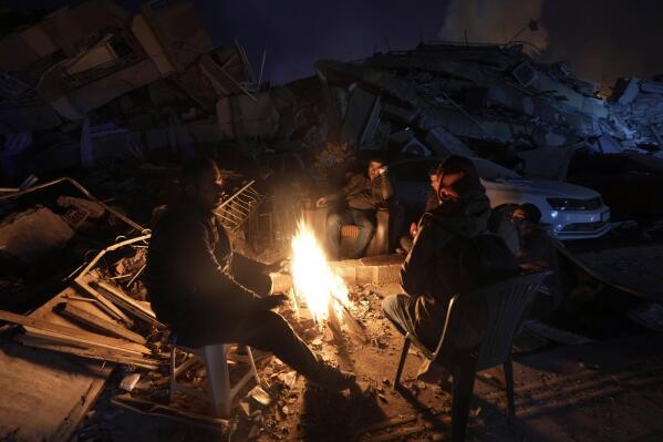 People warm up with fire in front of destroyed buildings in Antakya, southern Turkey, Wednesday, Feb. 8, 2023. With the hope of finding survivors fading, stretched rescue teams in Turkey and Syria searched Wednesday for signs of life in the rubble of thousands of buildings toppled by a catastrophic earthquake. (AP Photo/Khalil Hamra)