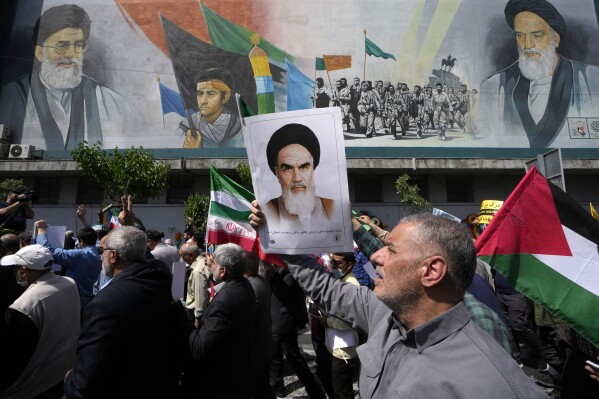 Iranian worshippers walk past a mural showing the late revolutionary founder Ayatollah Khomeini, right, Supreme Leader Ayatollah Ali Khamenei, left, and Basij paramilitary force, as they hold posters of Ayatollah Khomeini and Iranian and Palestinian flags in an anti-Israeli gathering after Friday prayers in Tehran, Iran, Friday, April 19, 2024. An apparent Israeli drone attack on Iran saw troops fire air defenses at a major air base and a nuclear site early Friday morning near the central city of Isfahan, an assault coming in retaliation for Tehran's unprecedented drone-and-missile assault on the country. (AP Photo/Vahid Salemi)