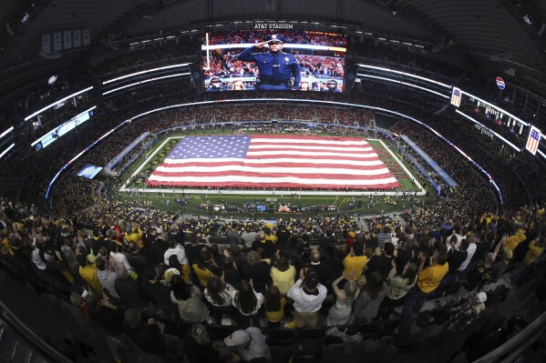 Fans at AT&T Stadium stand as a large U.S. flag is displayed on the field during the national anthem before the Cotton Bowl NCAA college football game between Ohio State and Missouri on Friday, Dec. 29, 2023, in Arlington, Texas. (AP Photo/Richard W. Rodriguez)