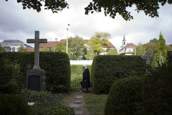 In this Sept. 18, 2019, photo, Sister Brigitte Queisser of the Lutheran Lazarus Order stands near graves of the Lazarus sisters during an interview with The Associated Press in Berlin. For many years, the deaconesses lived in West Berlin across the street from the Berlin Wall in a mother house that is seen in the background. The sisters’ order ran a hospital where many of the eastern escapees were treated. (AP Photo/Markus Schreiber)