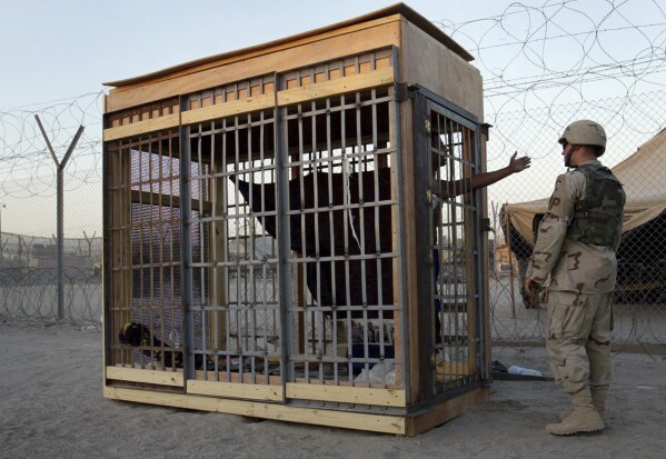 FILE - In this June 22, 2004, photo, a detainee in an outdoor solitary confinement cell talks with a military police officer at the Abu Ghraib prison on the outskirts of Baghdad, Iraq. A trial scheduled to begin Monday, April 15, 2024, in U.S. District Court in Alexandria, Va., will be the first time that survivors of Iraq’s Abu Ghraib prison will bring their claims of torture to a U.S. jury. Twenty years earlier, photos of abused prisoners and smiling U.S. soldiers guarding them shocked the world. (AP Photo/John Moore, File)