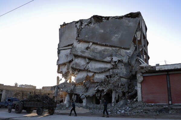 People walk next to the damage of a building that was destroyed in the February 2023 earthquake, in Idlib province, Syria, Saturday, Jan. 27, 2024. A year after the devastating 7.8 magnitude earthquake struck southern Turkey and northwestern Syria, a massive rebuilding effort is still trudging along. The quake caused widespread destruction and the loss of over 59,000 lives. (AP Photo/Omar Albam)