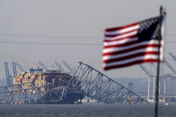 Dinsdag 26 maart 2024, Pasadena, MD.  (AP Photo/Mark Schiefelbein) Een Amerikaanse vlag wappert op een afmeerschip terwijl het containerschip Dolly tegen het wrak van de Francis Scott Key Bridge rust, hier te zien.