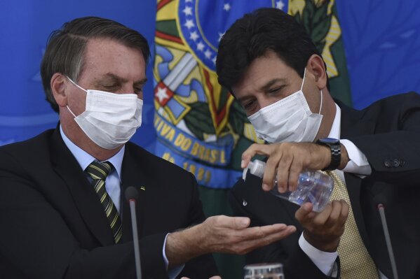 FILE - In this March 18, 2019 file photo, Brazil's Health Minister Luiz Henrique Mandetta, right, gives anti-bacterial gel to President Jair Bolsonaro as they give a press conference on the new coronavirus at Planalto presidential palace in Brasilia, Brazil. Mandetta criticized Bolsonaro’s dismissive handling of the COVID-19 pandemic on national television Sunday night, April 12, and the president’s repeated threats to fire him are worrying health experts who say that amid governmental chaos, the health minister's advice to limit contact and take the virus seriously has played a major role in preventing Brazil’s epidemic from being even worse.  (AP Photo/Andre Borges, File )