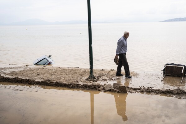 A man walks on the seaside past a car that was washed away by flood water in the town of Agria near the city of Volos, Greece, Thursday, Sept. 28, 2023. Torrential rainfall has caused widespread flooding in and around the Greek city of Volos and other central areas of the country for the second time in less than a month. (AP Photo/Petros Giannakouris)