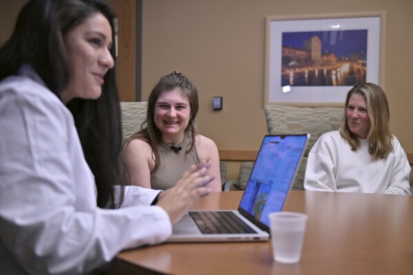 Alexis Bogan, center, and her mother Pamela Bogan, right, react to hearing a recreation of her lost voice from a prompt typed by Dr. Fatima Mirza, left, on Thursday, March 11, 2024, at Rhode Island Hospital in Providence, R.I. Doctors treating Bogan, who's speech was impaired by a brain tumor, used a voice-cloning tool from OpenAI to recreate her previous voice. (AP Photo/Josh Reynolds)