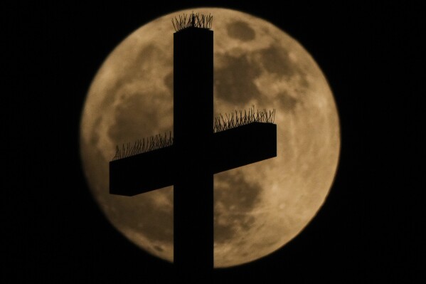 A cross atop the Cathedral of the Immaculate Conception Catholic Church is silhouetted against the rising full moon Sunday, May 11, 2025, in Kansas City, Mo. (AP Photo/Charlie Riedel)