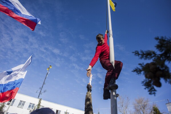 FILE - A member of a Pro-Russian self-defense force reaches for a knife, as he takes down a Ukrainian Navy flag at the Ukrainian Navy headquarters in Sevastopol, Crimea, Wednesday, March 19, 2014. The Crimean Peninsula's balmy beaches have been vacation spots for Russian czars and has hosted history-shaking meetings of world leaders. And it has been the site of ethnic persecutions, forced deportations and political repression. Now, as Russia’s war in Ukraine enters its 18th month, the Black Sea peninsula is again both a playground and a battleground. (AP Photo, File)