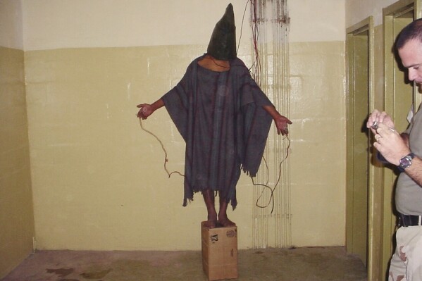FILE - This late 2003 photo obtained by The Associated Press shows an unidentified detainee standing on a box with a bag on his head and wires attached to him in the Abu Ghraib prison in Baghdad, Iraq. A trial scheduled to begin Monday, April 15, 2024, in U.S. District Court in Alexandria, Va., will be the first time that survivors of Iraq’s Abu Ghraib prison will bring their claims of torture to a U.S. jury. Twenty years ago, photos of abused prisoners and smiling U.S. soldiers guarding them shocked the world. (AP Photo, File)