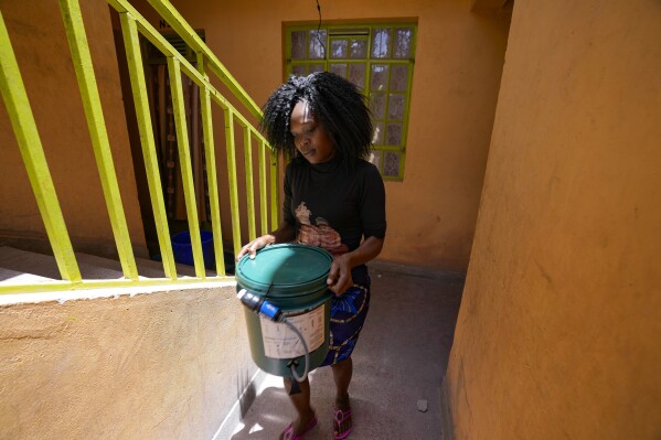 Meda Wekesa carries her water bucket filter to her house in Athi River, Machakos county, Kenya, Tuesday, Oct. 17, 2023. The devices are the size of a small water bottle and are fitted with a hose pipe onto a bucket. They can filter water from the river and nearby swamps into potable water that can be used by residents. (AP Photo/Brian Inganga)