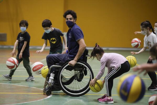Adolfo Damian Berdun, of Argentina, a professional player and captain of the Argentine basketball Paralympic team, teaches children basketball at a primary school in Verano Brianza, on the outskirts of Milan, Italy, Tuesday, May 11, 2021. Four second-grade classes in the Milan suburb of Verano Brianza have been learning to play basketball this spring from a real pro. They also getting a lesson in diversity.  Their basketball coach for the last month has been Adolfo Damian Berdun, an Argentinian-Italian wheelchair basketball champion. Berdun, 39, lost his left leg in a traffic accident at ag 13 in his native Buenos Aires, and he has visited many schools over the years to discuss how he has lived with his disability. (AP Photo/Luca Bruno)