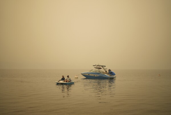 Smoke fills the air as Pat Manzuik and her husband Trevor use a paddleboat to get to shore after being given a boat ride by good samaritan Christy Dewalt, back right, back to their home they were evacuated from due to the Lower East Adams Lake wildfire, in Scotch Creek, British Columbia, Sunday, Aug. 20, 2023. (Darryl Dyck/The Canadian Press via AP)