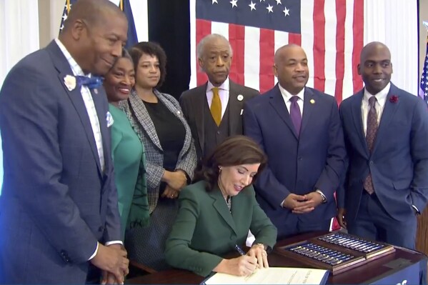 CORRECTS CITY TO NEW YORK - This image from video provided by the Office of The Governor shows New York Gov. Kathy Hochul signing a bill in New York, Tuesday, Dec. 19, 2023, to create a commission tasked with considering reparations to address the persistent, harmful effects of slavery in the state. She is joined by, standing from left: Dr. Yohuru Williams, Founding Director of the Racial Justice Initiative at the University of St. Thomas; Andrea Stewart-Cousins, Majority Leader of the NY State Senate; Michaelle Solages, NY State Assembly Woman; Rev. Al Sharpton; Carl Heastie, Speaker of the NY State Assembly; James Sanders, NY State Senator. (Office of the Governor via AP)