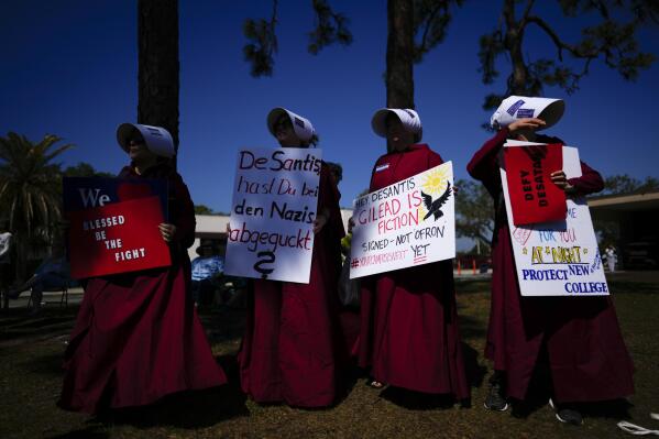 FILE -Joyce White, center left, whose daughter Lola is a third-year biology major at New College of Florida, protests along with other parents dressed as handmaids from Margaret Atwood's "The Handmaid's Tale," ahead of a meeting by the college's board of trustees, Tuesday, Feb. 28, 2023, on the school's campus in Sarasota, Fla. White, carries a sign in German addressed to Florida Governor Ron DeSantis that reads, "DeSantis, Are you copying the Nazis?" For parents who have felt the intense heartache of watching their child struggle and then the deep relief of seeing them thrive, the upheaval at New College is making them relive a painful past.(AP Photo/Rebecca Blackwell, File)