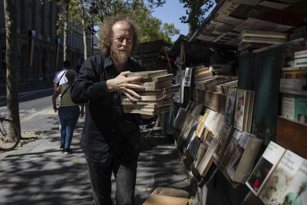 Jerome Callais, head of the Paris Booksellers' Cultural Association, carries books at his bookseller booth along the Seine Riverbank in Paris, Tuesday, Aug. 22, 2023. The host city of Paris vowed to deliver an extraordinary grand opening on July 26, 2024, as the ceremony is expected to draw about 600,000 spectators to the Parisian quayside. Citing security measures, the Paris police prefecture ordered on July 25 the removal of 570 stationary boxes out of which booksellers have operated for decades on the quays of the Seine river. (AP Photo/Sophie Garcia)