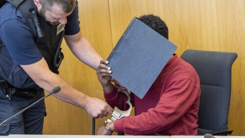 A court official, left, removes the handcuffs from the defendant, who is covering his face with a file folder, in the hearing room of the Regional Court in Ulm, Germany, Tuesday, July 4, 2023. A court in Germany has convicted a man who attacked two girls and killed one of them last year and sentenced him to life in prison. (Stefan Puchner/dpa via AP)