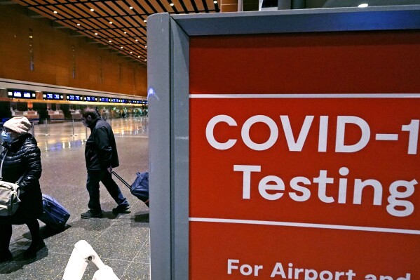 FILE - Travelers pass a sign near a COVID-19 testing site in Terminal E at Logan Airport, on Dec. 21, 2021, in Boston. The nation’s top public health agency is expanding a program that tests international travelers for COVID-19 and other infectious diseases. The Centers for Disease Control and Prevention currently operates a program at six U.S. airports that asks passengers from inbound international flights to agree to nose swabs and answer questions about their travel. (AP Photo/Charles Krupa, File)