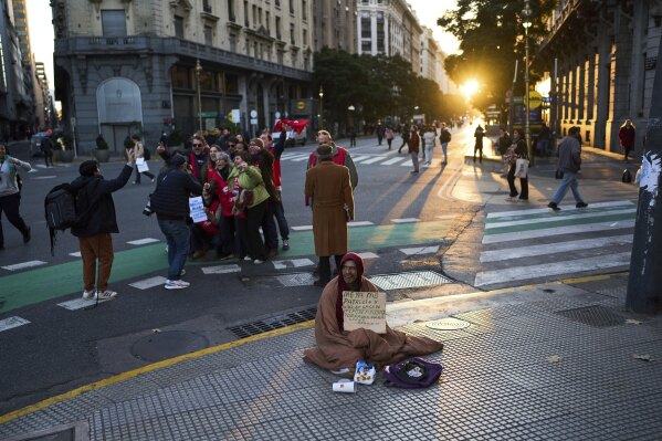 Patricio Roblez, que sostiene un cartel en el que explica que no tiene casa, pide limosna mientras manifestantes antigubernamentales posan para una selfie en grupo, en Buenos Aires, Argentina, el 11 de junio de 2025. (AP Foto/Rodrigo Abd)