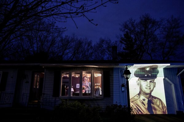 An image of veteran Francis Foley is projected onto the home of his wife, Dale Foley, left, as she looks out a window with their daughter, Keri Rutherford, in Chicopee, Mass., Wednesday, April 29, 2020. Foley, a U.S. Army veteran and resident of the Soldier's Home in Holyoke, Mass., died from the COVID-19 virus at the age of 84. Seeking to capture moments of private mourning at a time of global isolation, the photographer used a projector to cast large images of veterans on to the homes as their loved ones are struggling to honor them during a lockdown that has sidelined many funeral traditions. (AP Photo/David Goldman)