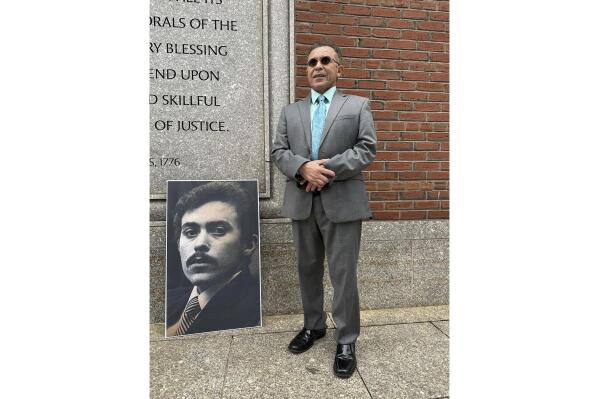 Victor Rosario, 65, stands next to a photo of himself at age 24 when he was on trial in 1982 for arson, Wednesday, May 3, 2023, outside the federal courthouse in Boston. Rosario, who spent 32 years in prison after he was wrongfully convicted of setting a fire that killed eight people, will receive $13 million from the city of Lowell, Mass., in a settlement of a lawsuit he brought against the city in 2019. (AP Photo/Mark Pratt)