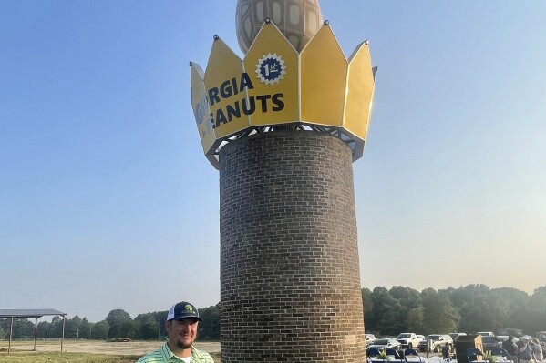 This photo provided by the Georgia Peanut Commission shows Cole Sercer of Sercer Machine & Fabrication, standing beside the Big Peanut that he and his employees designed and built near Interstate 75 in Ashburn, Ga., Thursday, July 20, 2023. The monument replaces a similar peanut that blew down during Hurricane Michael in October 2018. (Joy Crosby/Georgia Peanut Commission via AP)