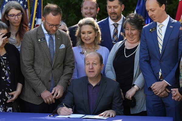 Colorado Gov. Jared Polis, center, prepares to sign as bill sponsors Sen. Dylan Roberts, far right, and Rep. Matt Soper, far left, look on to bring more comprehensive guidelines into place for funeral facilities Friday, May 24, 2024, during a ceremony outside the Governor's mansion in downtown Denver. (AP Photo/David Zalubowski)