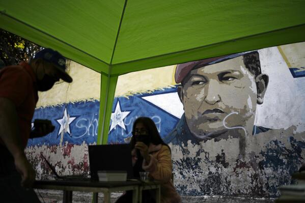 A mural of late President Hugo Chavez decorates a street wall where a table is set up, by the National Election Council (CNE), where people can sign a petition in favor of holding a referendum to remove President Nicolas Maduro from office in Caracas, Venezuela, Wednesday, Jan. 26, 2022. The signatures of 20 percent of registered voter must be collected within 12 hours to request a presidential recall, a CNE rule that Maduro's opposition criticizes as impossible. (AP Photo/Matias Delacroix)