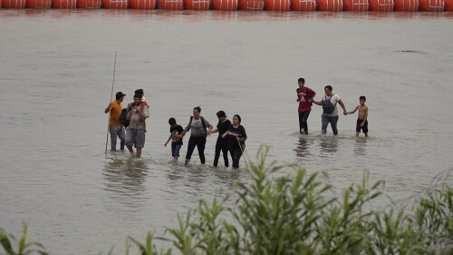 Migrants who crossed the Rio Grande from Mexico walk past large buoys being deployed as a border barrier on the river in Eagle Pass, Texas, Wednesday, July 12, 2023. The floating barrier is being deployed in an effort to block migrants from entering Texas from Mexico. (AP Photo/Eric Gay)