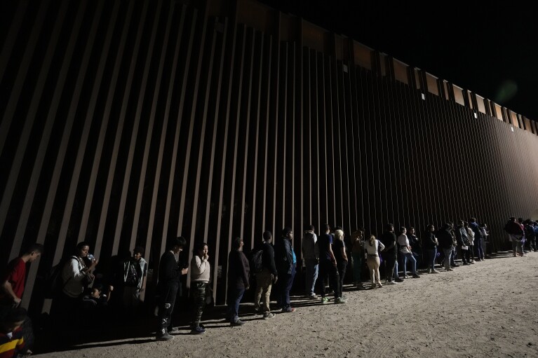 FILE - People line up against a border wall as they wait to apply for asylum after crossing the border from Mexico Tuesday, July 11, 2023, near Yuma, Arizona. In Senegal's capital, Nicaragua is a hot ticket among travel agents serving people who want to live in the United States. Many migrants take various flights to eventually arrive there legally and then journey illegally by land to the U.S. border with Mexico. (AP Photo/Gregory Bull)