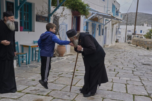 O monge cristão ortodoxo padre Spyridon de Amorgos beija a mão do filho de seu sobrinho Nikitas Vasalos enquanto o padre Kostas observa, na vila de Lagada, ilha de Amorgos, Grécia, no sábado, 29 de março de 2025. (Foto AP/Petros Giannakouris)