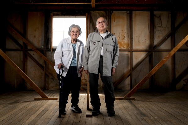 Minidoka survivor Paul Tomita, right, poses for a photograph with his wife Mabel Imai Tomita, a Gila River and Tule Lake survivor, in an original barracks at Minidoka National Historic Site, Saturday, July 8, 2023, in Jerome, Idaho. Paul, who had asthma as an infant, remembers the desert dust repeatedly sending him to the camp hospital. Now in their 80s, the Tomitas have traveled to participate in many pilgrimages to Japanese American incarceration sites. "We have to make sure that it doesn't happen again. And the only way we do is we have to participate," said Paul Tomita. (AP Photo/Lindsey Wasson)