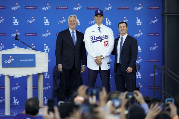 Los Angeles Dodgers' Shohei Ohtani, center, poses for a photo with owner and chairman Mark Walter, left, and president of baseball operations Andrew Friedman applaud during a news conference at Dodger Stadium Thursday, Dec. 14, 2023, in Los Angeles. (AP Photo/Ashley Landis)