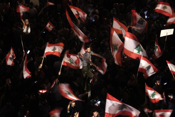 FILE - In this Oct. 21, 2019 file photo, anti-government protesters wave Lebanese flags and chant,  "the people want to bring down the regime" outside the office of legislator Mohammed Raad, the po...