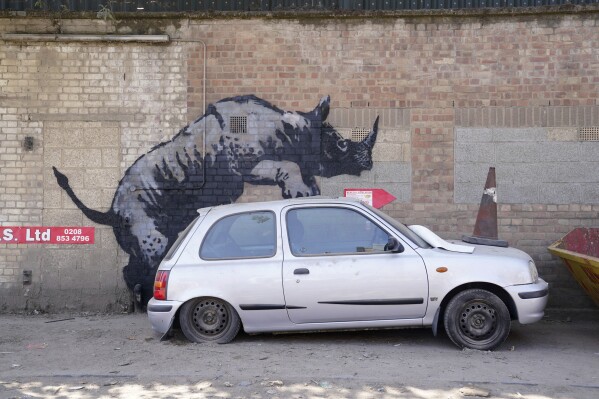 A new artwork unveiled by Banksy, depicting a rhinoceros which looks as though it is climbing on top of a car, the eighth artwork in his animal-themed collection, on Westmoor Street in Charlton, south east London, Monday, Aug. 12, 2024. (Lucy North/PA via AP)