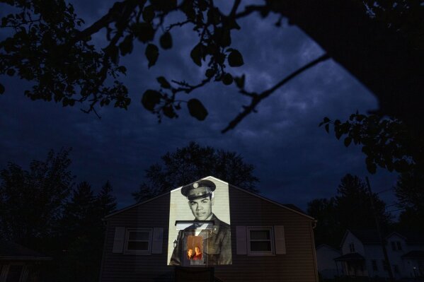 An image of veteran Samuel Melendez is projected onto the home of his nieces, Janet Ramirez, right, and Mary Perez, as they look out a doorway in Chicopee, Mass., Sunday, May 17, 2020. Originally of Puerto Rico, Melendez, a U.S. Army Korean War veteran and resident of the Soldier's Home in Holyoke, Mass., died from the COVID-19 virus at the age of 86. Seeking to capture moments of private mourning at a time of global isolation, the photographer used a projector to cast large images of veterans on to the homes as their loved ones are struggling to honor them during a lockdown that has sidelined many funeral traditions. (AP Photo/David Goldman)