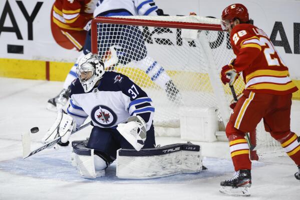 Winnipeg Jets goalie Connor Hellebuyck, left, watches the puck as Calgary Flames' Elias Lindholm looks on during second-period NHL hockey game action in Calgary, Alberta, Monday, Feb. 21, 2022. (Jeff McIntosh/The Canadian Press via AP)