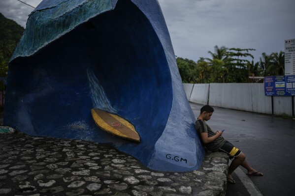 A resident sits by a sculpture of the Teahupo'o wave at the end of the road, Tahiti, French Polynesia, Saturday, Jan. 13, 2024. (AP Photo/Daniel Cole)