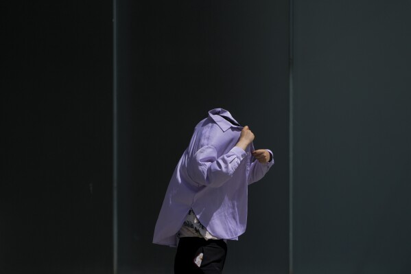 FILE - A woman uses a shirt to shield from the sun as she walks at an outdoor shopping mall on a sweltering day in Beijing, July 6, 2023. Climate change is making heat waves crawl slower across the globe and last longer with higher temperatures over larger areas, a new study finds. (AP Photo/Andy Wong, File)