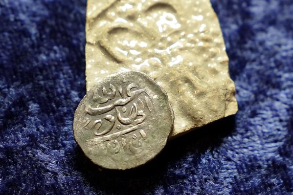A 17th century Arabian silver coin that research shows was struck in 1693 in Yemen, rests against a piece of 17th century broken pottery featuring a likeness of Queen Mary, on a table in Warwick, R.I., Thursday, March 11, 2021. The coin was found at a farm, in Middletown, R.I., in 2014 by metal detectorist Jim Bailey, who contends it was plundered by English pirate Henry Every in 1695 from Muslim pilgrims sailing home to India after a pilgrimage to Mecca. (AP Photo/Steven Senne)
