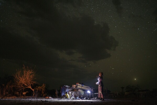 Amadou Altine Ndiaye holds a flashlight as he sits on his metal-framed bed next to his children and wife where they set up camp in the village of Yawara Dieri, in the Matam region of Senegal, Saturday, April 15, 2023. In Senegal, caravans carry the comforts of a furnished home, such as a metal bed frame and mattress, and water for people and animals. (AP Photo/Leo Correa)