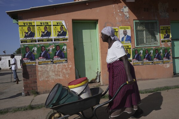 An elderly woman pushes a wheelbarrow past campaign posters in Mabvuku on the outskirts of Harare, Saturday, Dec. 9, 2023. Zimbabwe was holding by-elections Saturday for nine seats in Parliament after opposition lawmakers were removed from their positions and disqualified from standing again in what they have called an illegal push by the ruling ZANU-PF party to bolster its majority and have the power to change the constitution. (AP Photo/Tsvangirayi Mukwazhi)